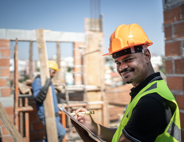 Profissional de obra sorrindo, utilizando capacete de proteção e segurando projetos de construção.