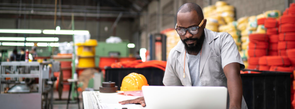 Dono de loja de material de construção trabalha em seu computador no estoque da empresa. Ele é um homem negro jovem que tem expressão de concentração