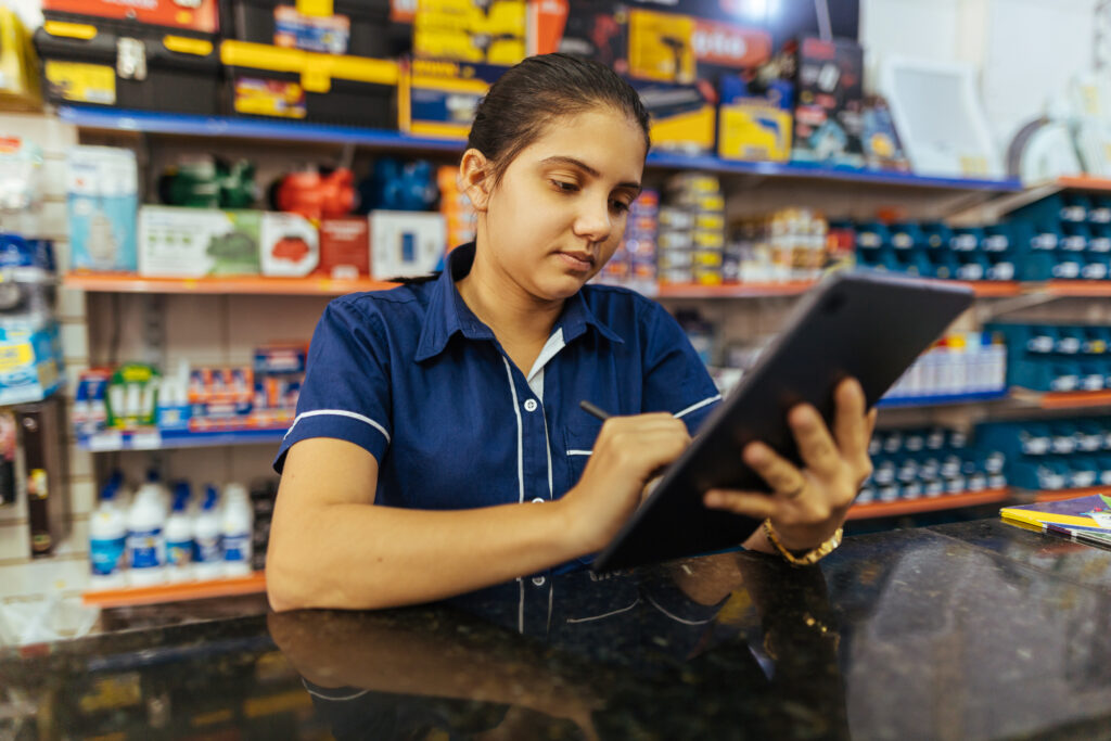 Gerente de loja de materiais de construção observa um tablet atenta. Ela é uma mulher latina jovem que está uniformizada atrás de um balcão.