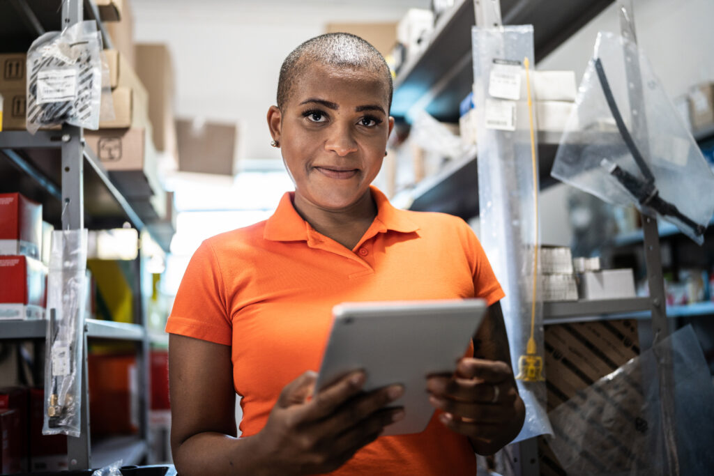 Foto de uma mulher fazendo inventário com um tablet em um estoque de materiais de construção. Ela é uma mulher negra jovem que está com olhar otimista e leve sorriso