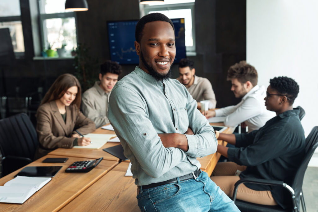 Jovem funcionário está em uma reunião, ele está posando para a câmera com um sorriso confiante e os braços cruzados. Ele é um homem negro com feições alegres.