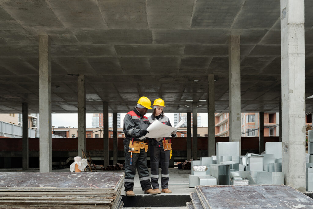 Dois profissionais de obra com roupas de trabalho e equipamentos de proteção individual (EPI) olham para o plano de construção. Eles são uma mulher branca e um homem negro, ambos são jovens e parecem concentrados.