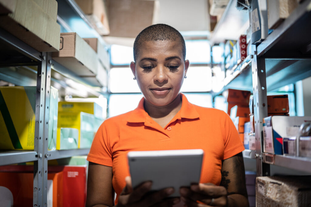 Mulher fazendo inventário para reposição do estoque com um tablet em uma loja de materiais de construção. Ela é negra e jovem, que está uniformizada e concentrada.