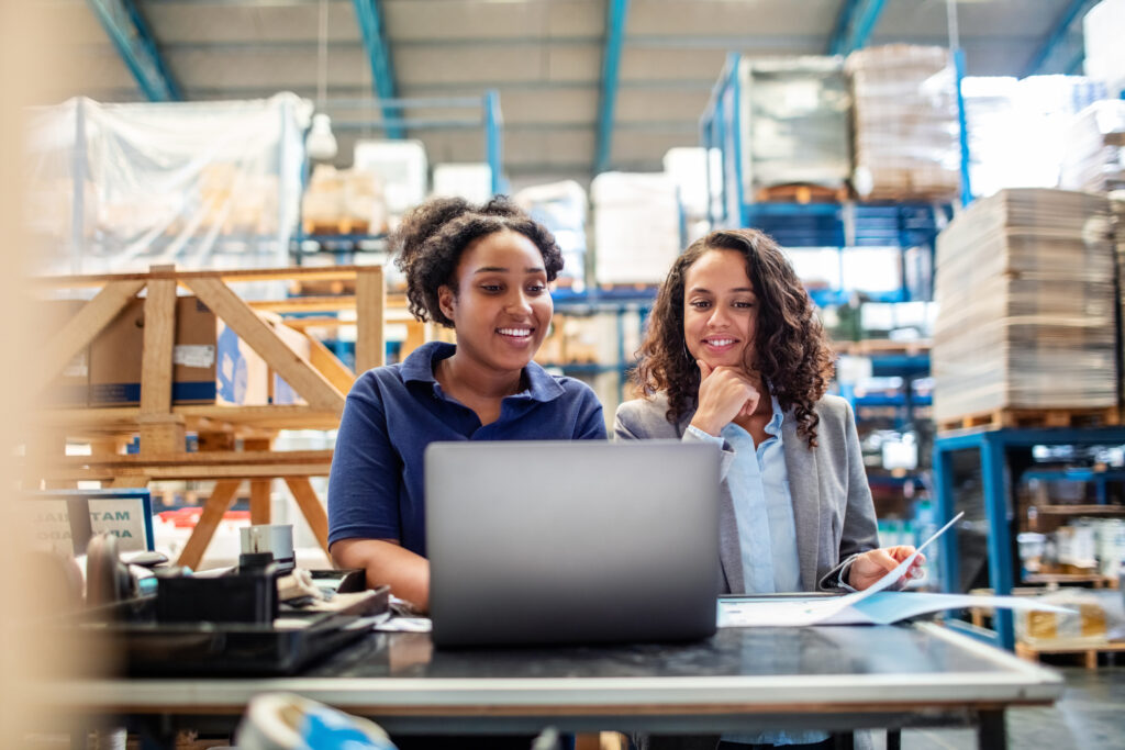 Duas mulheres conversam no estoque de uma loja de material de construção enquanto consultam um computador